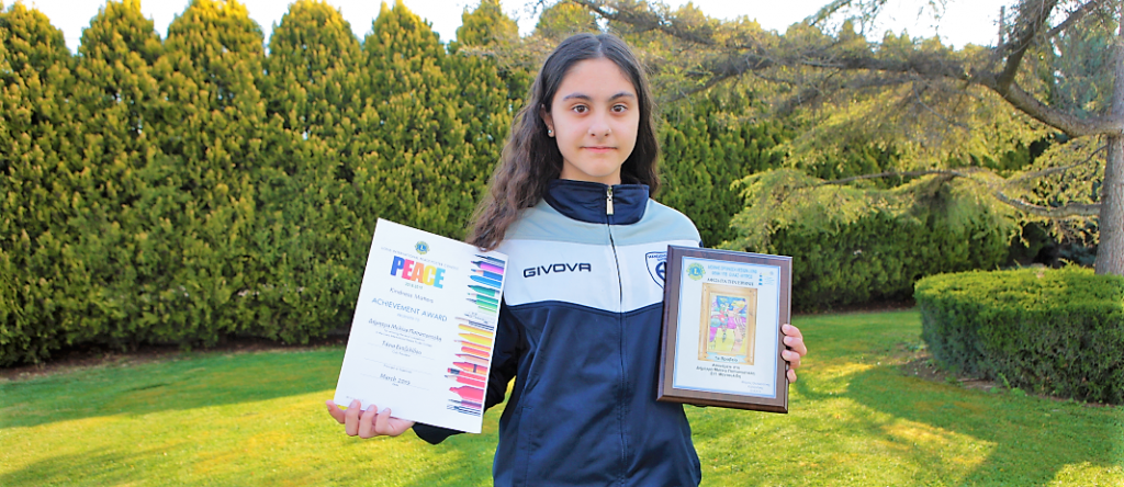 student D. - M. Papapostoli dressed in blue, holding the award she won, with green and trees behind her