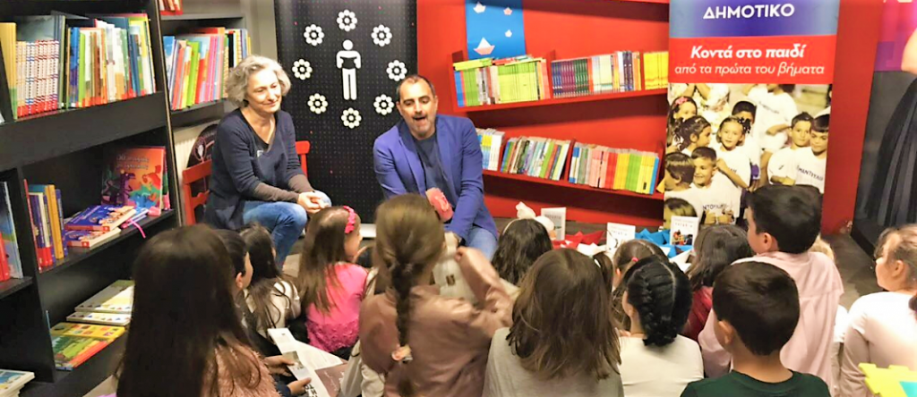Aris Dimokidis talks to the little students of mandoulides schools about his new book and the students sit down on the floor looking at him