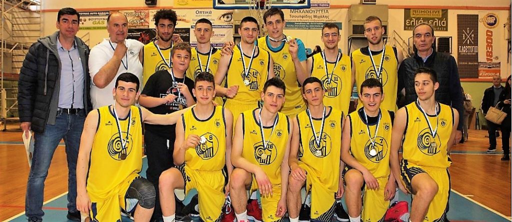 The boys of mandoulides basketball team wearign their yellow shirts, hugged with each other, 6 of them standing and 6 of them sitting, with their coaches near them