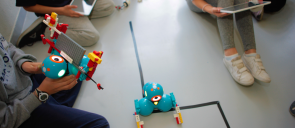 Ένα μπλέ ρομποτάκι στο κέντρο της εικόνας και τα παιδιά με tablets γύρω του