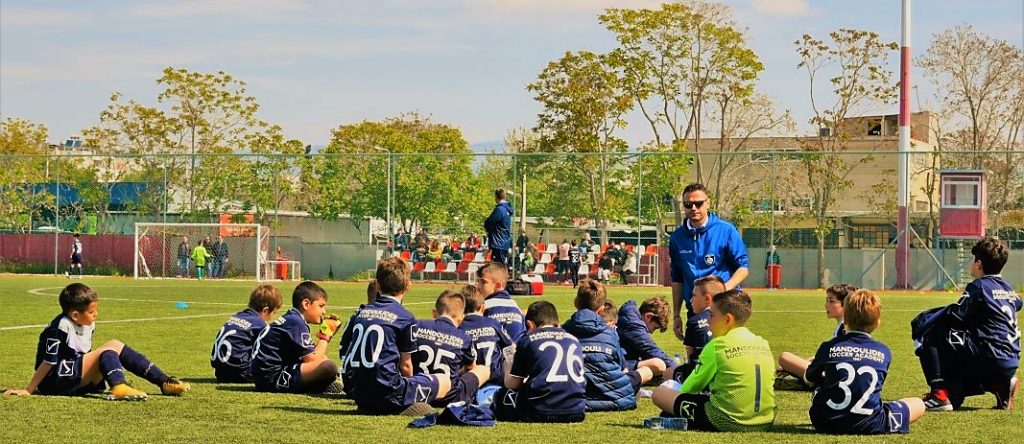 Στην Εκδρομή Ακαδημίας Ποδοσφαίρου στην Αθήνα, οι μικροί ποδοσφαιριστές των εκπαιδευτηρίων καθισμένοι στο γρασίδι του γηπέδου ποδοσφαίρου ακούν τον προπονητή τους