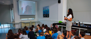Οι μαθητές του δημοτικού καθισμένοι στο πάτωμα μαζί με τη δασκάλα του όρθια δίπλα τους παρακολουθούν ομιλία μέσω skype
